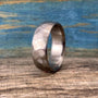 A silver-colored metal ring with a hammered exterior texture stands upright on a wooden surface against a blue backdrop. __etsy:8442154475