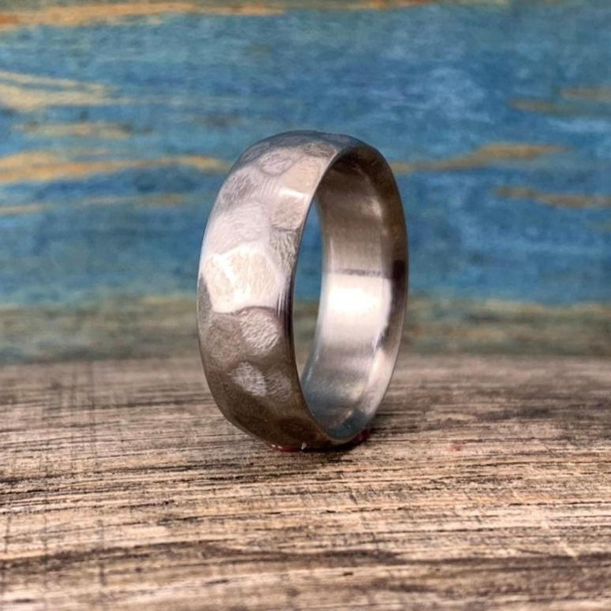 A silver-colored metal ring with a hammered exterior texture stands upright on a wooden surface against a blue backdrop. __etsy:8442154475