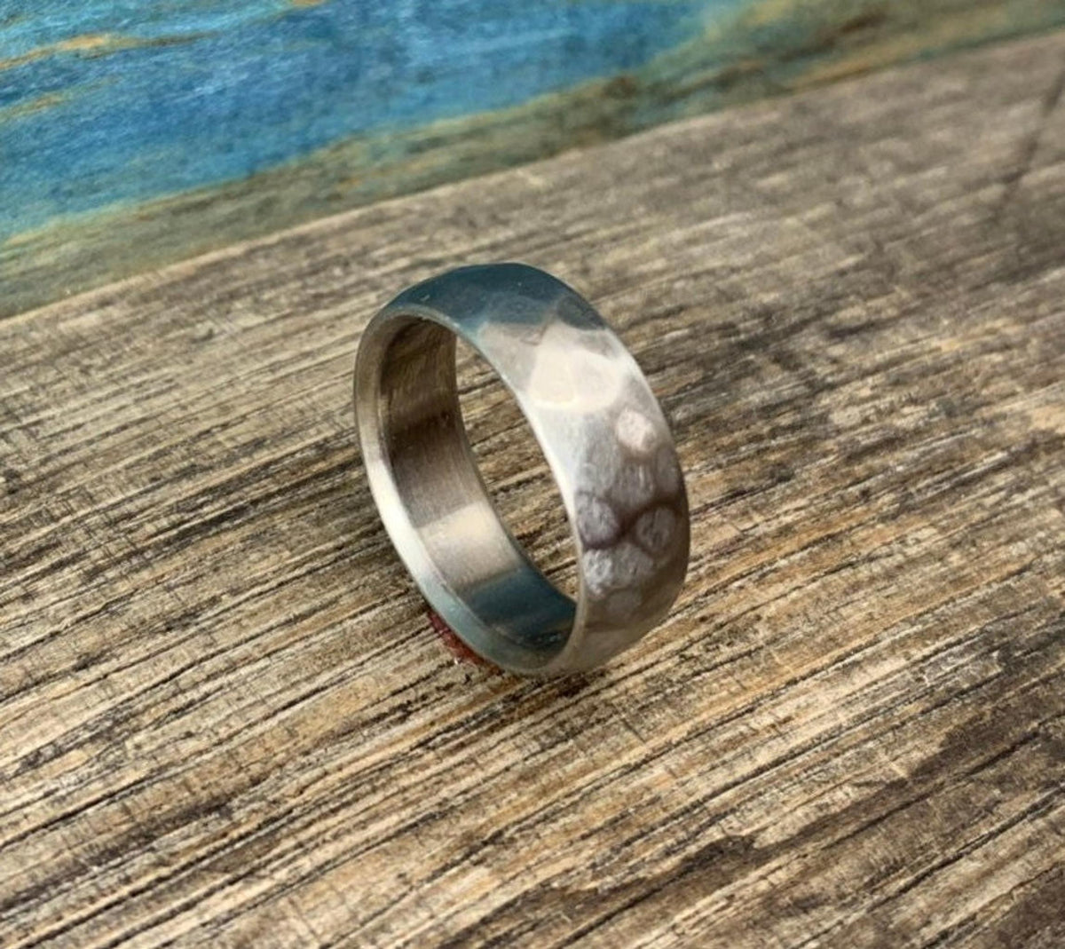 The textured metal ring stands upright diagonally on a rustic wooden surface with a blue background. __etsy:8394273678