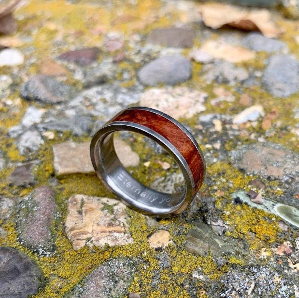 A silver-colored ring with a central band of reddish-brown wood, engraved with "Eternally yours" on the inside, rests on mossy rocks. __etsy:8381336602