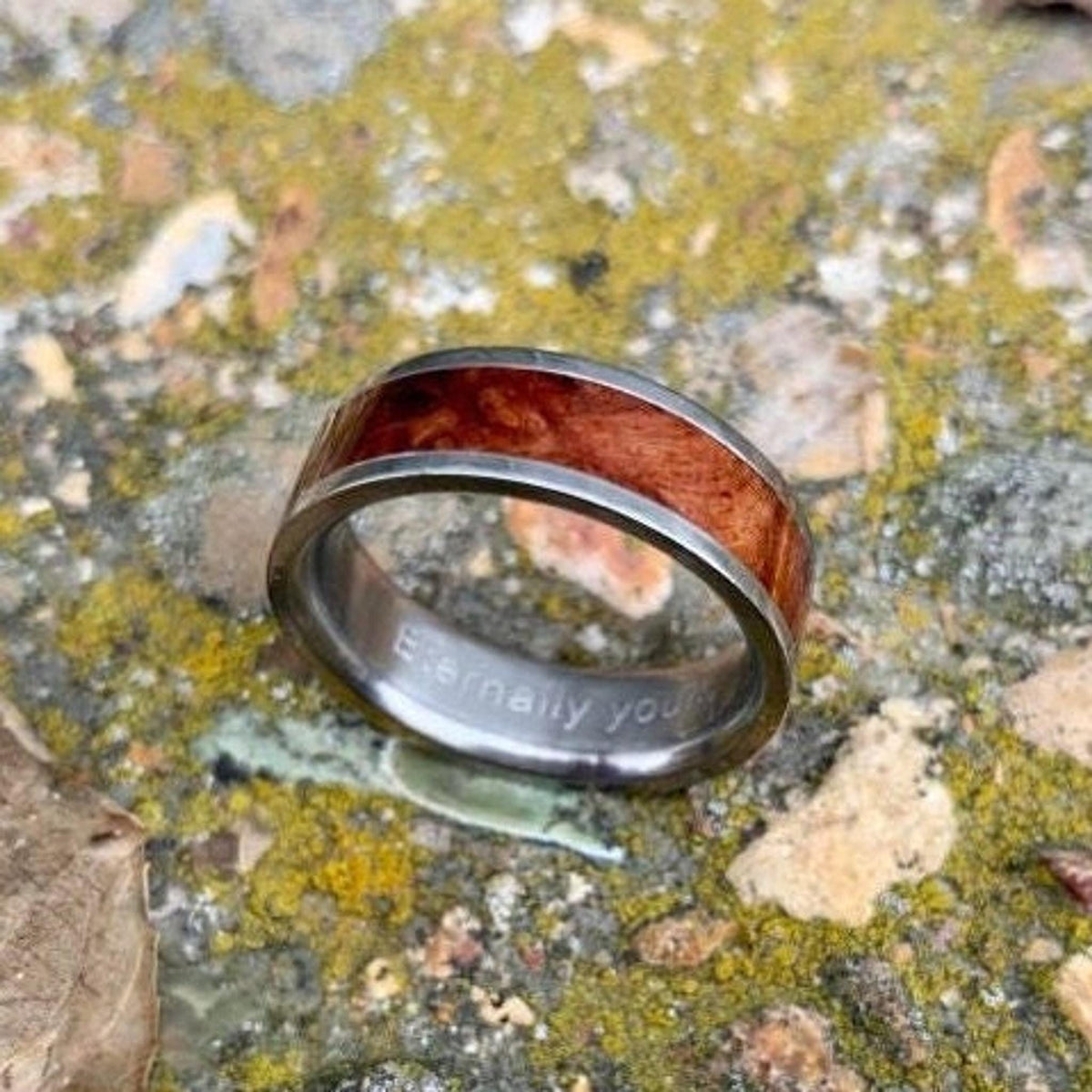 A silver-colored ring with a central band of reddish-brown wood, engraved with "Eternally yours" on the inside, rests on mossy ground. __etsy:8327914197