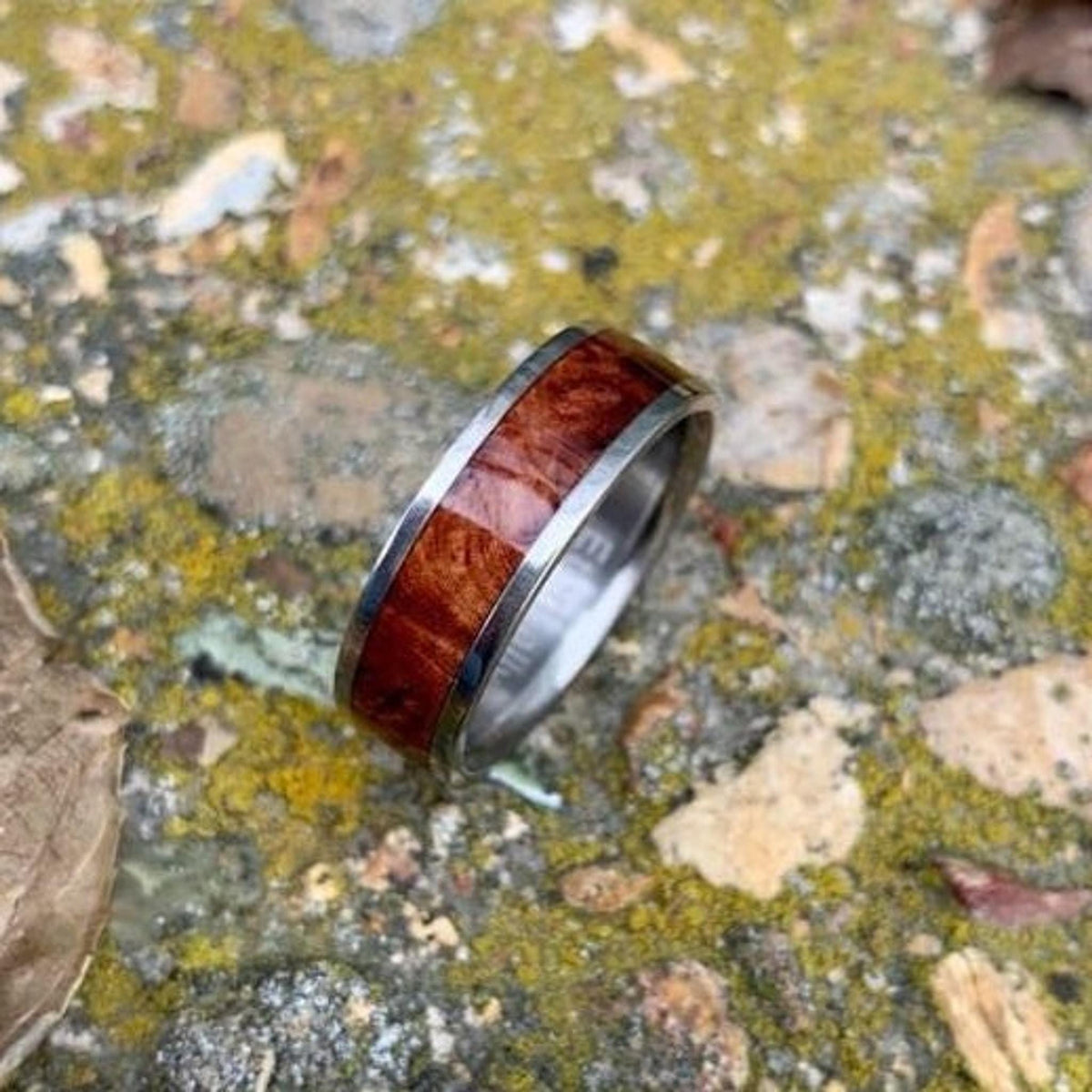 A silver-colored ring with a central band of reddish-brown wood is positioned on mossy ground with small stones. __etsy:8279994432