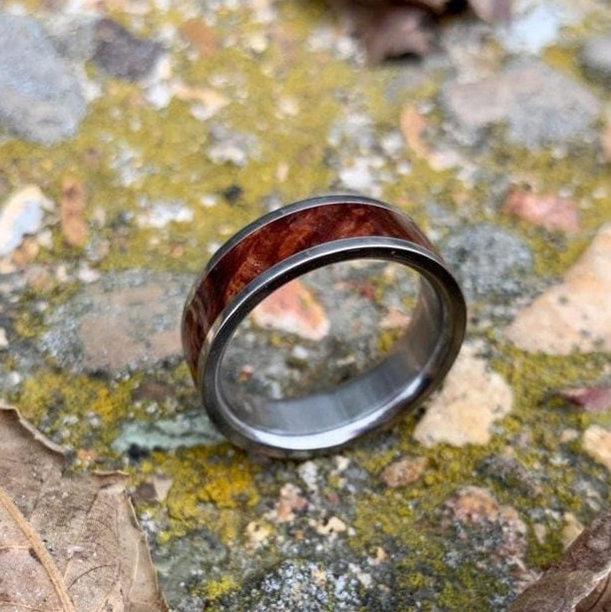 A silver-colored ring with a central band of reddish-brown wood is shown from a slight angle, resting on mossy ground and a dried leaf. __etsy:8327914639