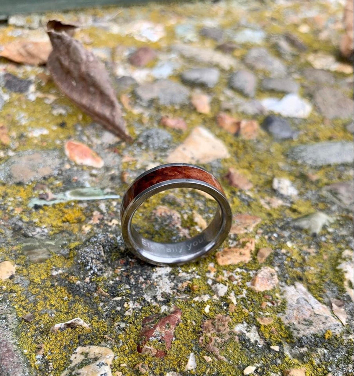 A silver-colored ring with a central band of reddish-brown wood, engraved with "Eternally yours" on the inside, is on mossy ground with a dried leaf in the background. __etsy:8327914883
