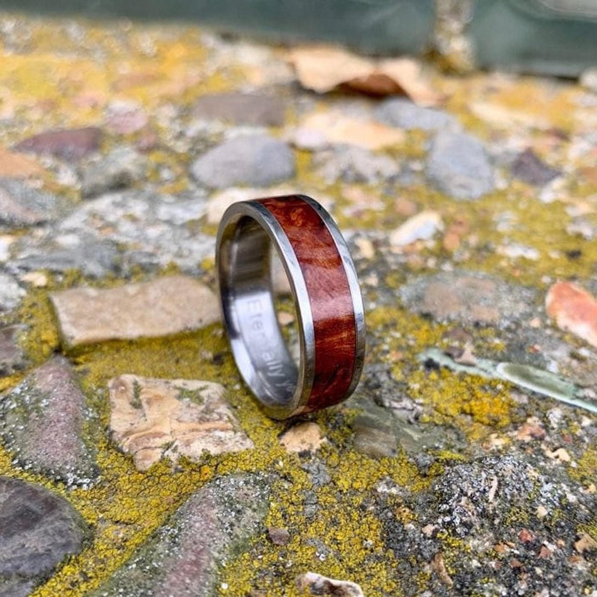 A silver-colored ring with a central band of reddish-brown wood, engraved with "Eternally" on the inside, rests on mossy rocks. __etsy:8327915045