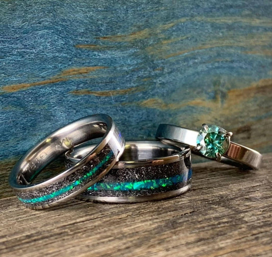 A wedding band set featuring two meteorite rings with bright teal opal inlays and a matching engagement ring with a teal moissanite stone resting on a rustic wooden surface. __etsy:8425545013