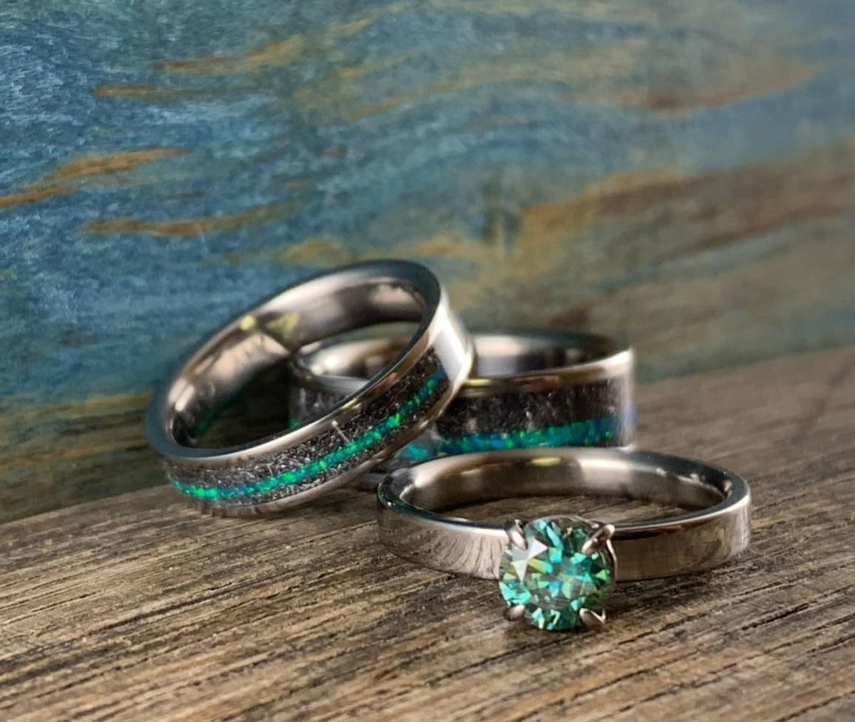 A teal moissanite solitaire ring placed in front of two meteorite wedding bands inlaid with teal opal. __etsy:8425545399