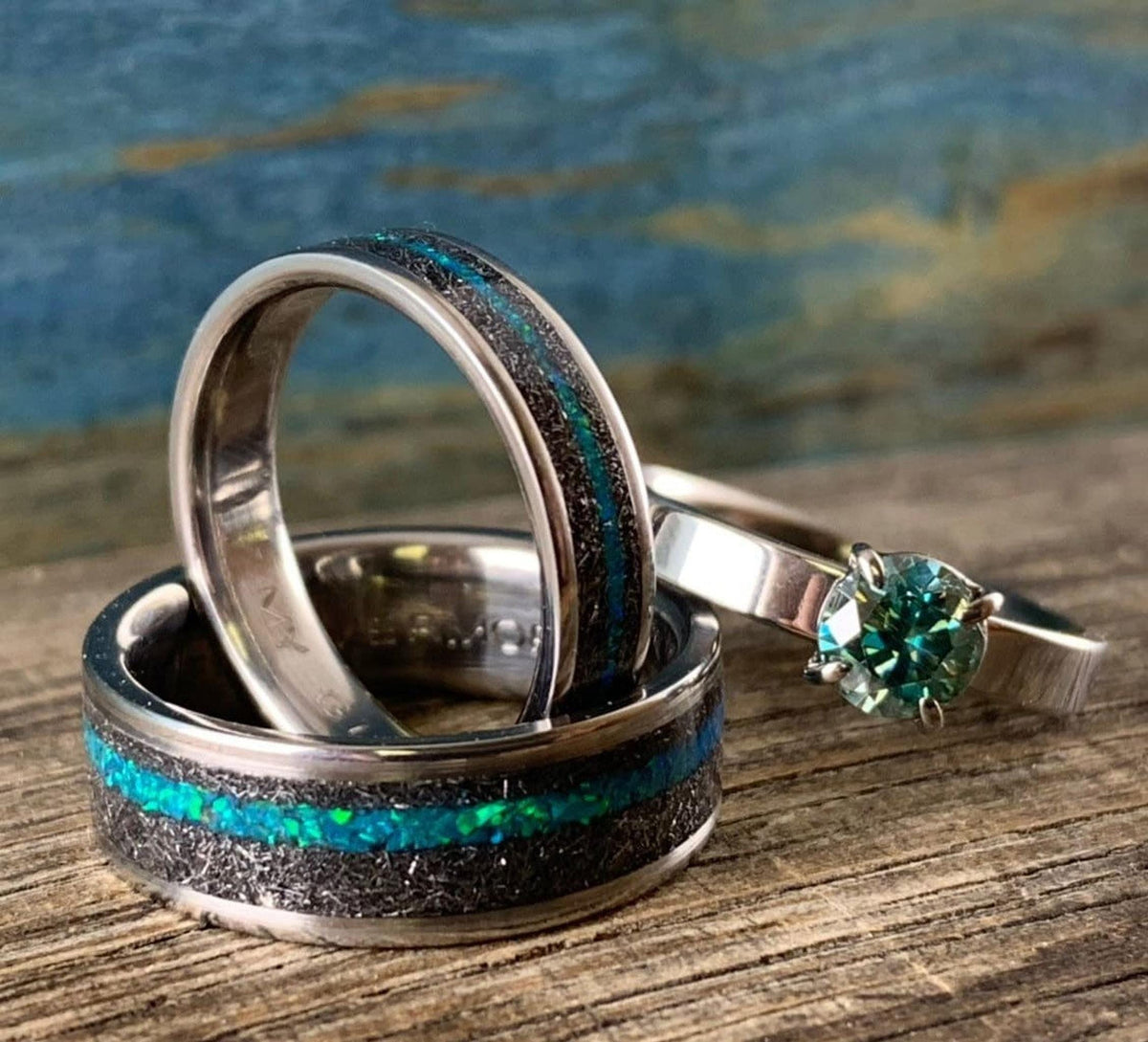 A wider meteorite and teal opal ring standing upright, leaning against a smaller matching band, with a teal moissanite ring positioned to the side. __etsy:8425545643