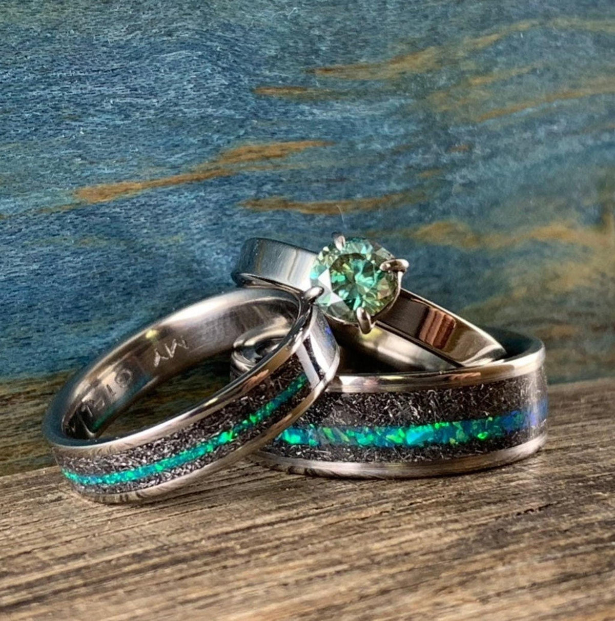 A teal moissanite engagement ring resting behind two meteorite bands featuring vibrant teal opal stripes. __etsy:8377671830