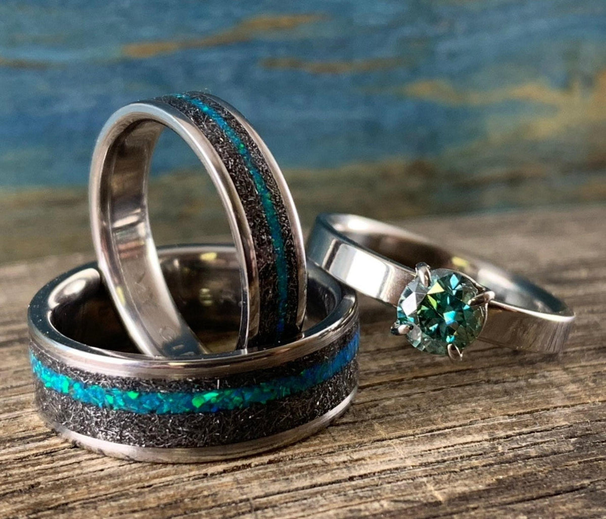 A wider meteorite band with a central teal opal inlay lying flat beneath an upright meteorite band and a teal moissanite engagement ring. __etsy:8377671942