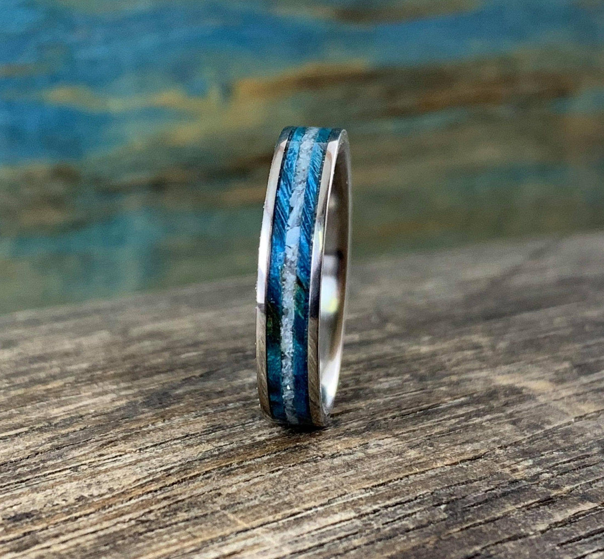 A silver-colored ring with a central inlay of blue wood and white pearl, viewed from a slight angle on a wooden surface.