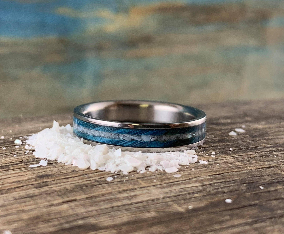 A silver-colored ring with a blue wood and white pearl inlay, sitting on a small pile of white crystalline flakes on a wooden surface.