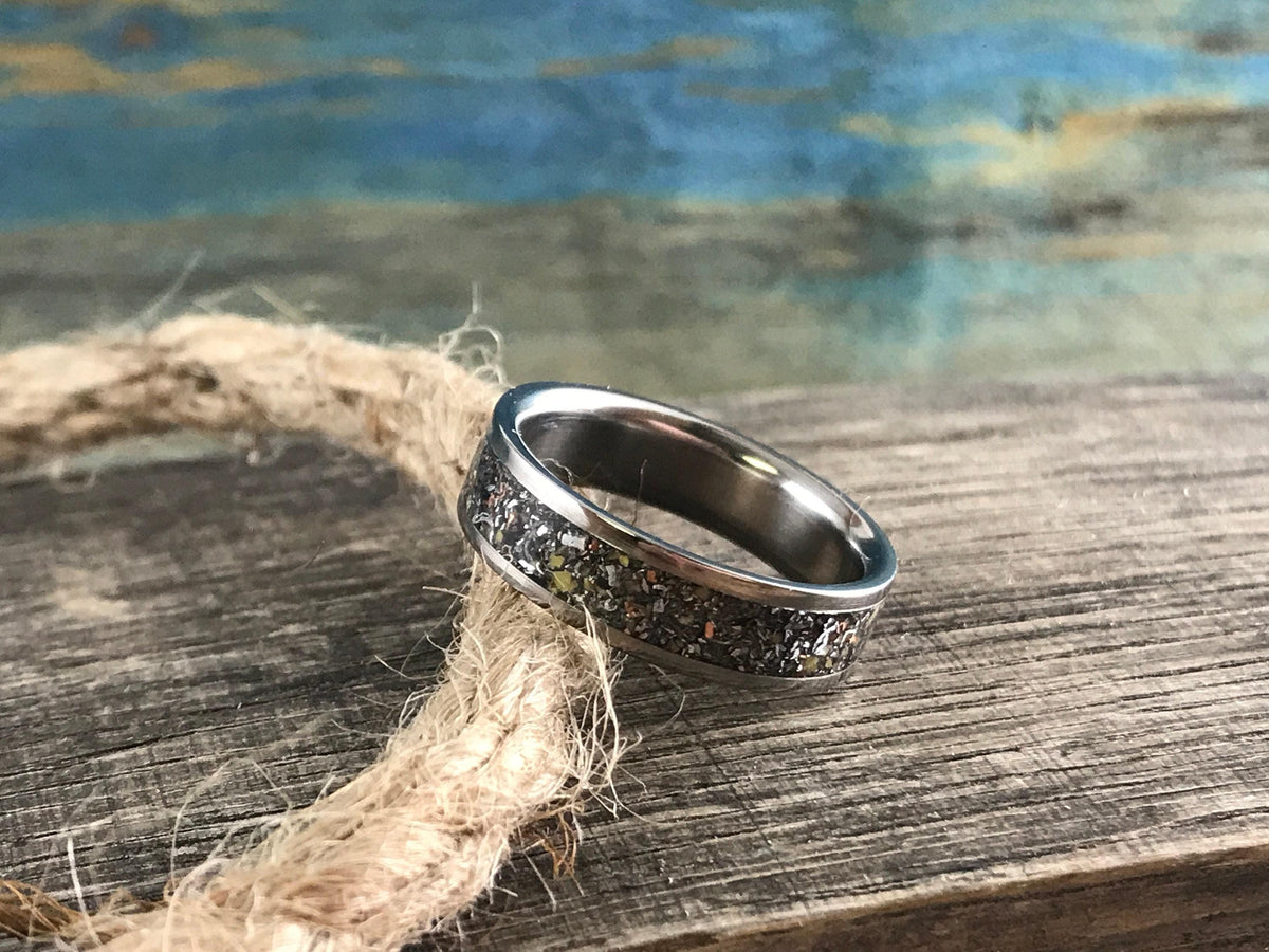 A silver-colored ring with a dark, textured inlay band is placed on a thick, braided rope, with a blue and brown background.