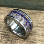 A silver-colored ring with a wide central band of purple and yellow flecks, bisected by a thin purple line, is shown at an angle on a wooden surface.
