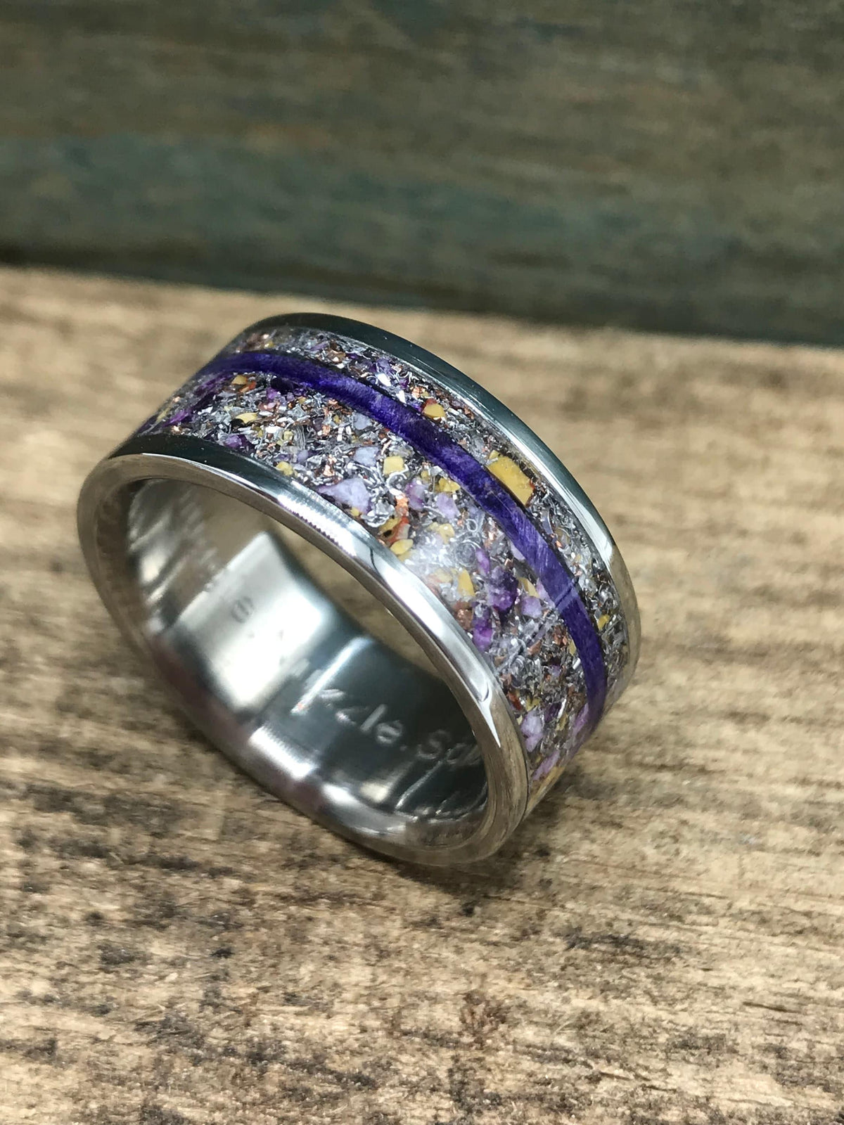 A silver-colored ring with a wide central band of purple and yellow flecks, bisected by a thin purple line, is shown at an angle on a wooden surface.