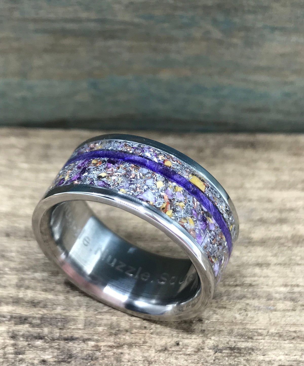 A silver-colored ring with a central inlay of purple and yellow flecks, divided by a thin purple stripe, is angled on a wooden surface.