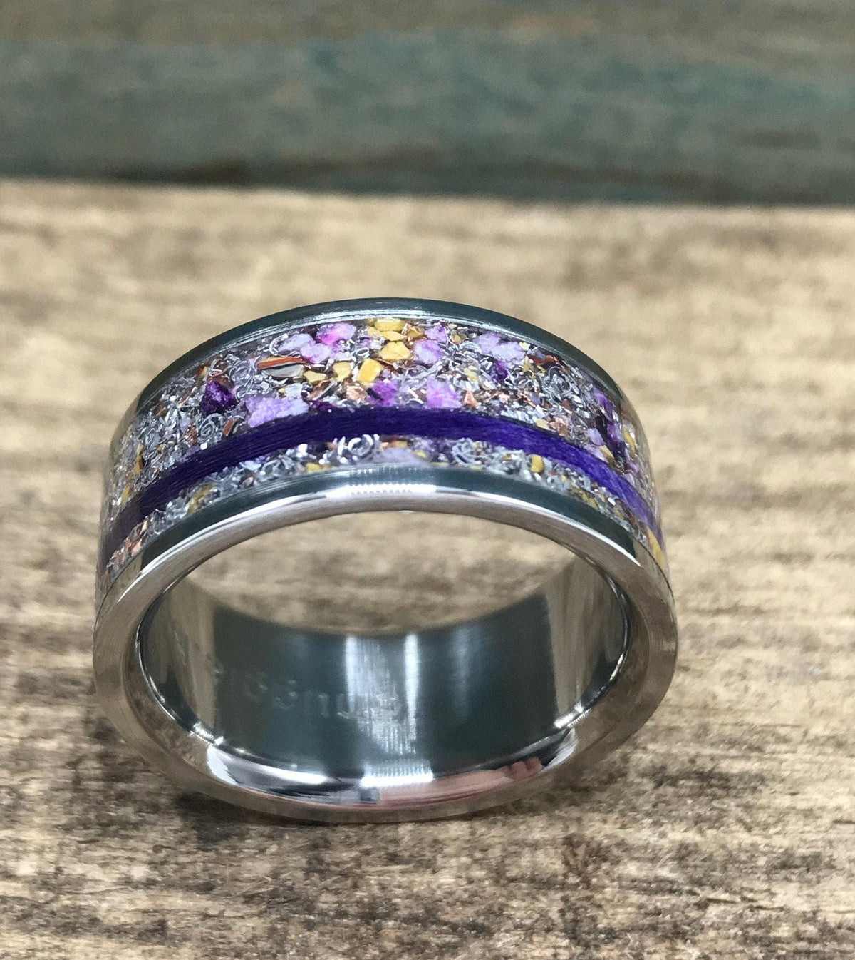 A silver-colored ring with a central inlay of purple and yellow flecks, bisected by a thin purple line, is shown head-on on a wooden surface.