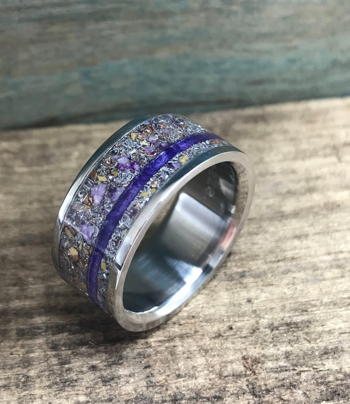 A silver-colored ring with a central band of purple and yellow flecks, bisected by a thin purple line, is shown at an angle on a wooden surface.