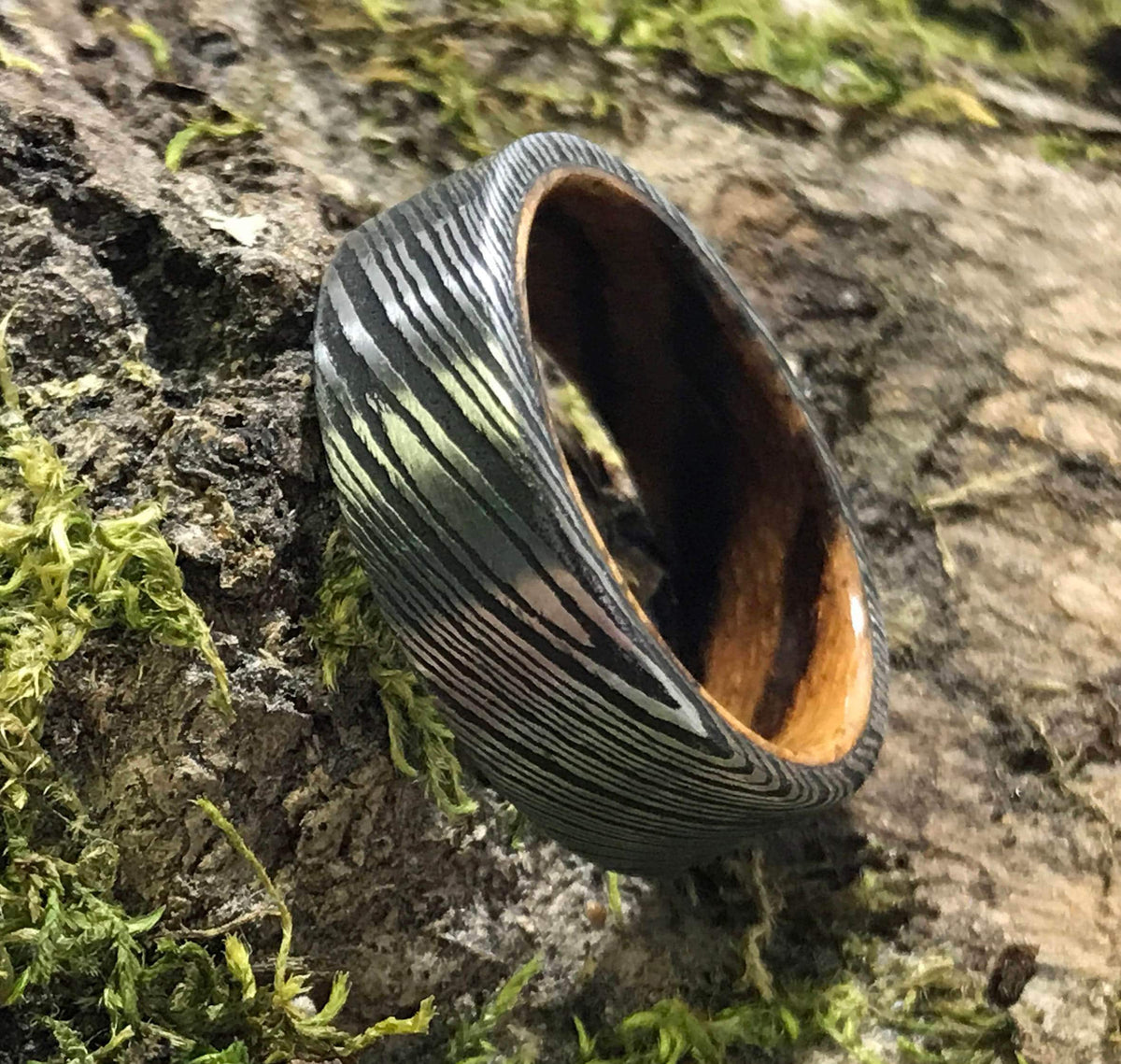 A Damascus steel ring with a zebrawood interior is placed on a piece of bark covered in green moss, viewed from an angle.