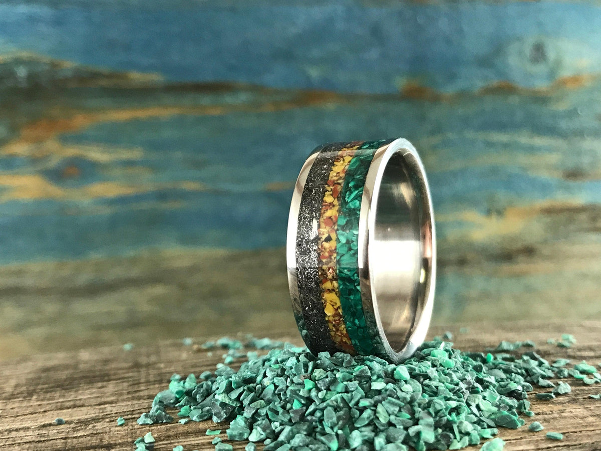A silver ring with green, yellow, and black inlay bands stands upright on a pile of small green malachite chips.