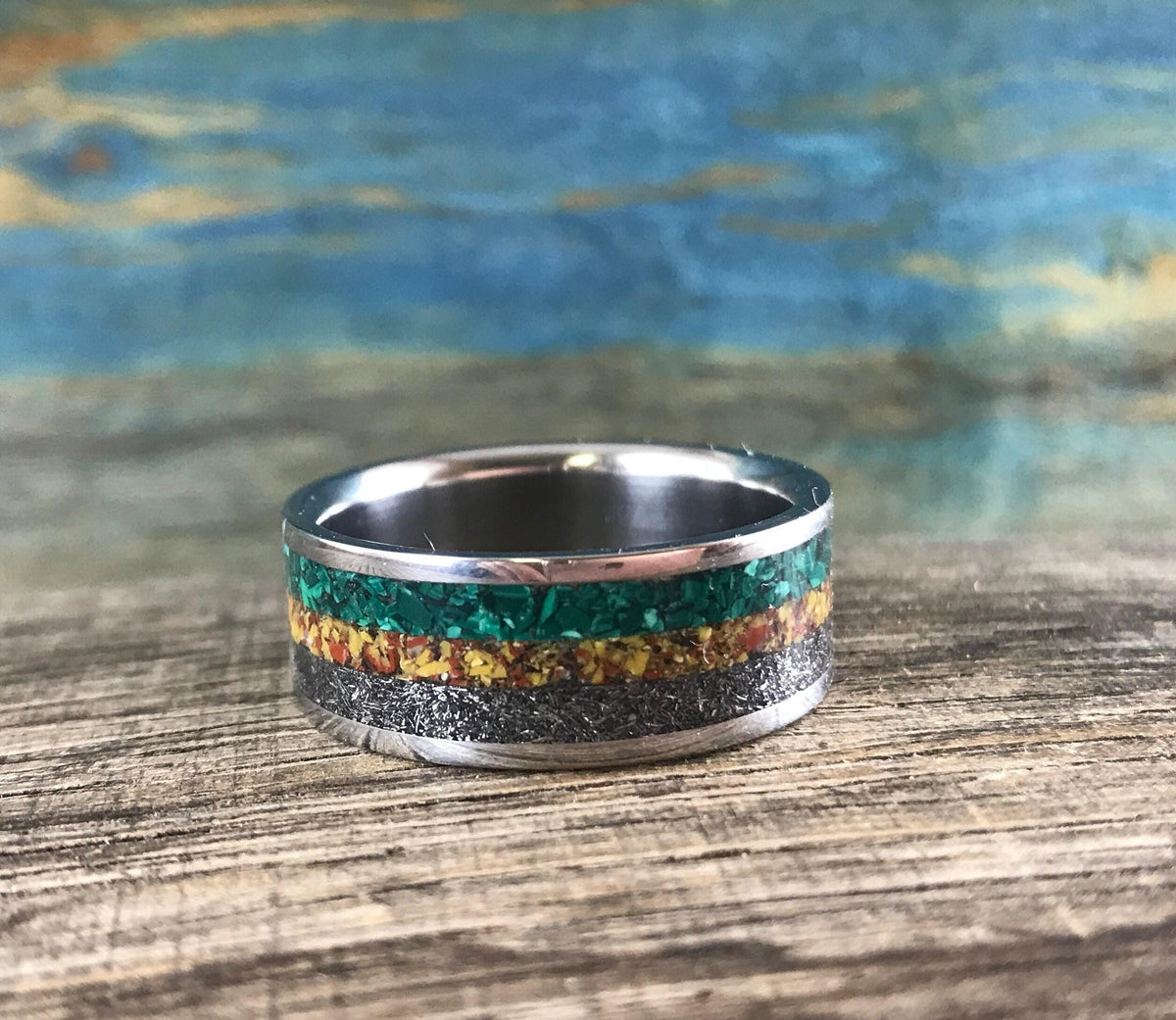 A silver ring with green, yellow, and black inlay bands is shown from the front on a light brown wooden surface.