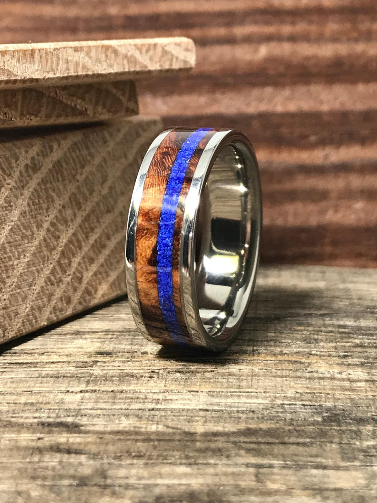 A silver-colored ring with a central band of brown wood and a thin blue stripe, resting on a wooden surface.