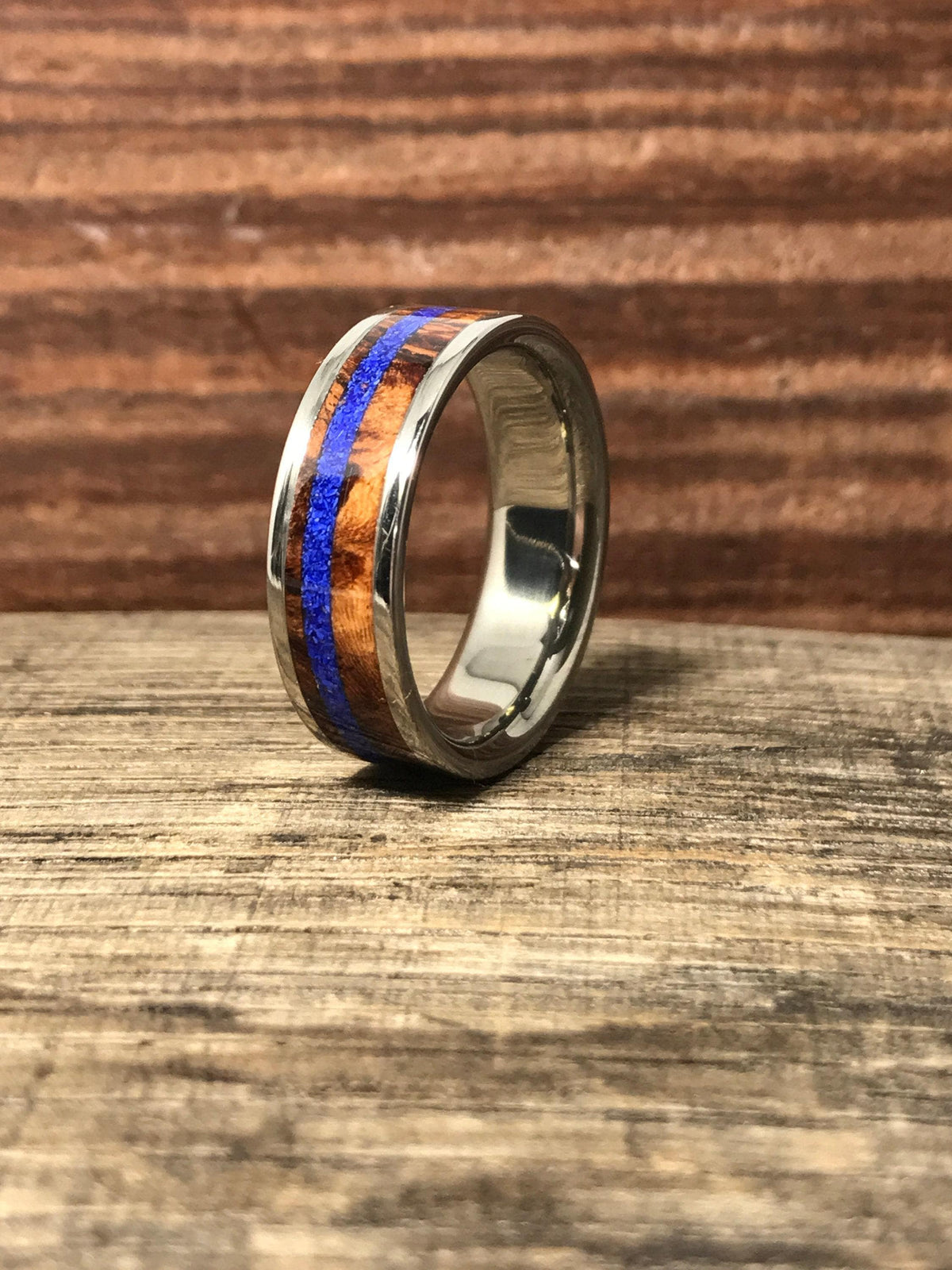 A silver-colored ring with a central brown wood band and a thin blue stripe, centered on a wooden surface.