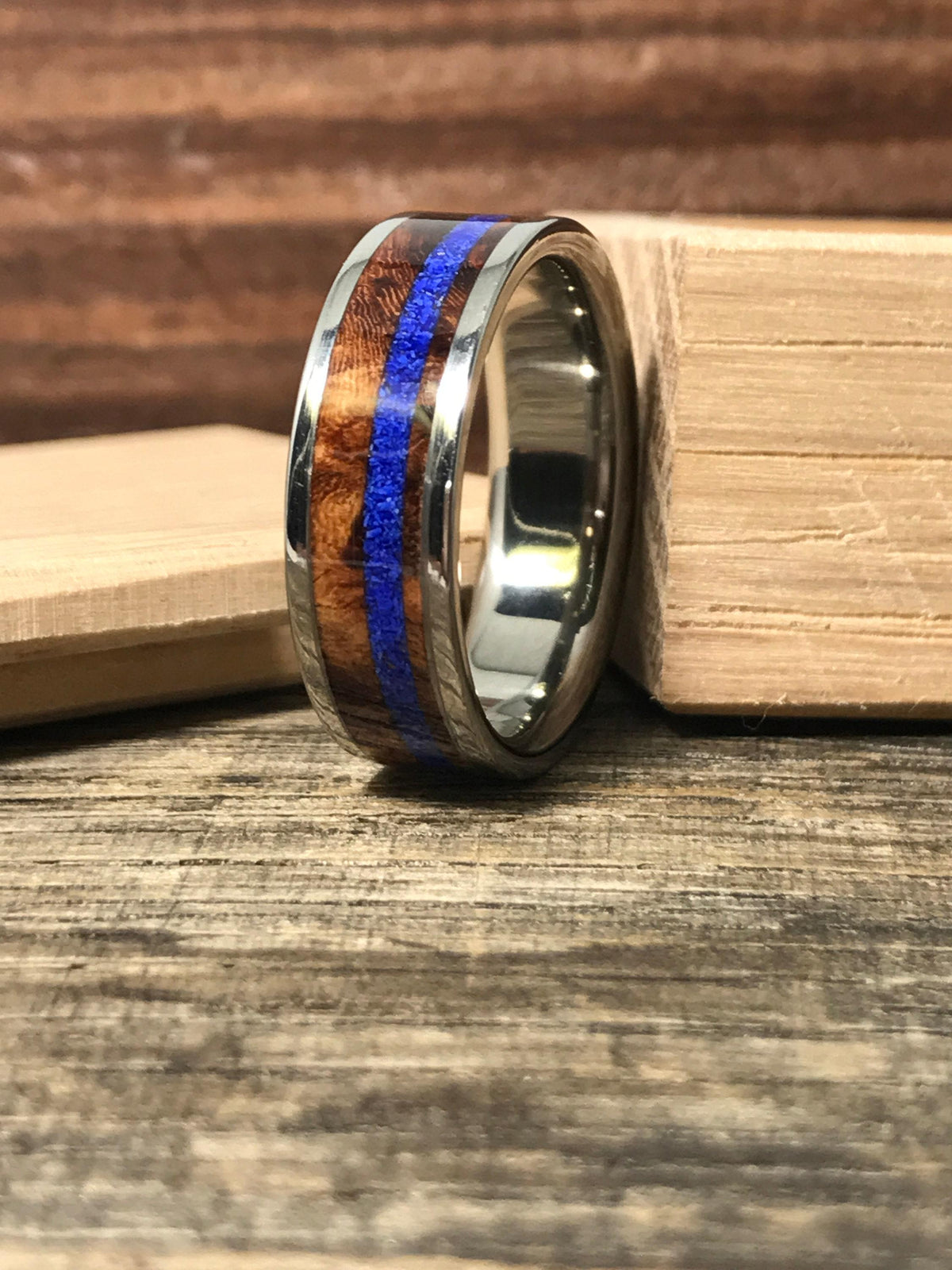 A silver-colored ring with a central brown wood band and a thin blue stripe, resting on a wooden surface next to a light wood block.