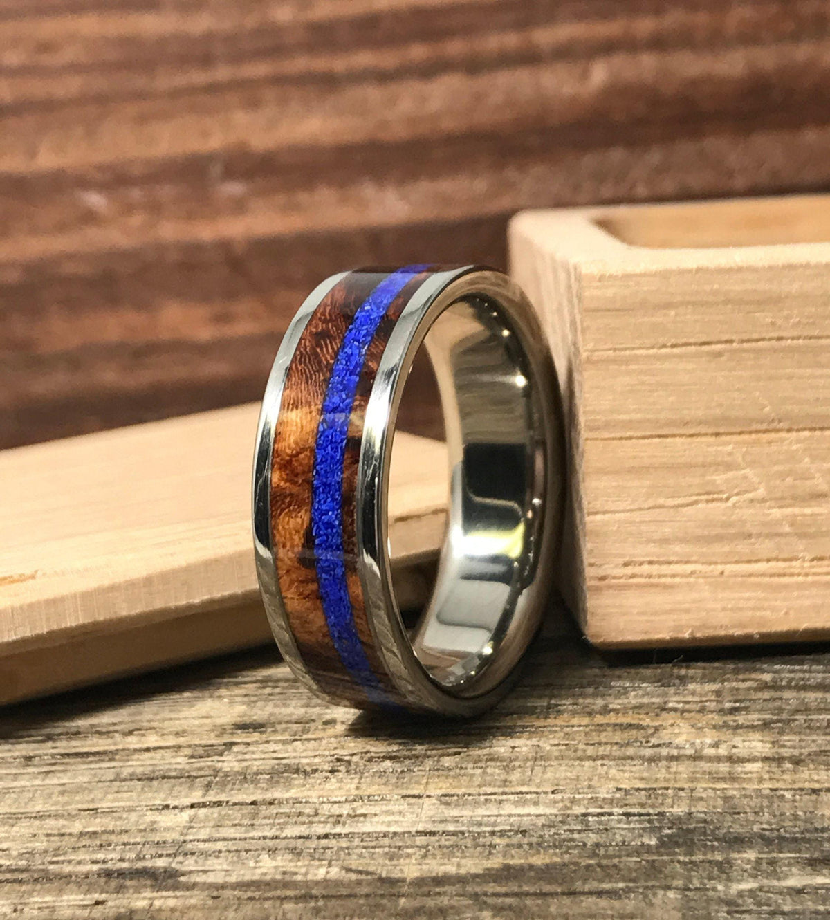 A silver-colored ring with a central brown wood band and a thin blue stripe, resting on a wooden surface with a light wood box in the background.