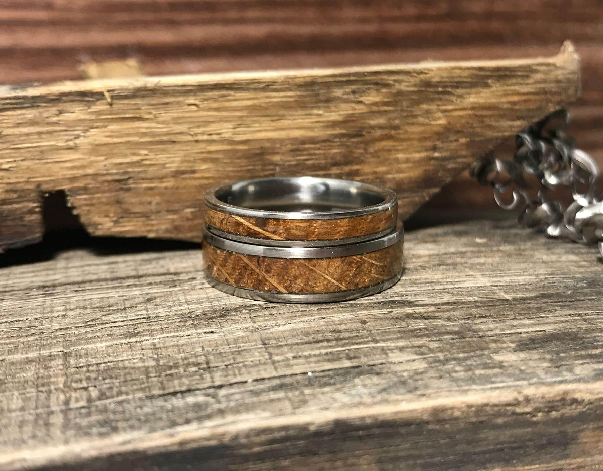 A single silver-toned ring with a light brown wood inlay is centered on a weathered wooden surface, with a piece of rough-cut wood and a silver chain in the background.