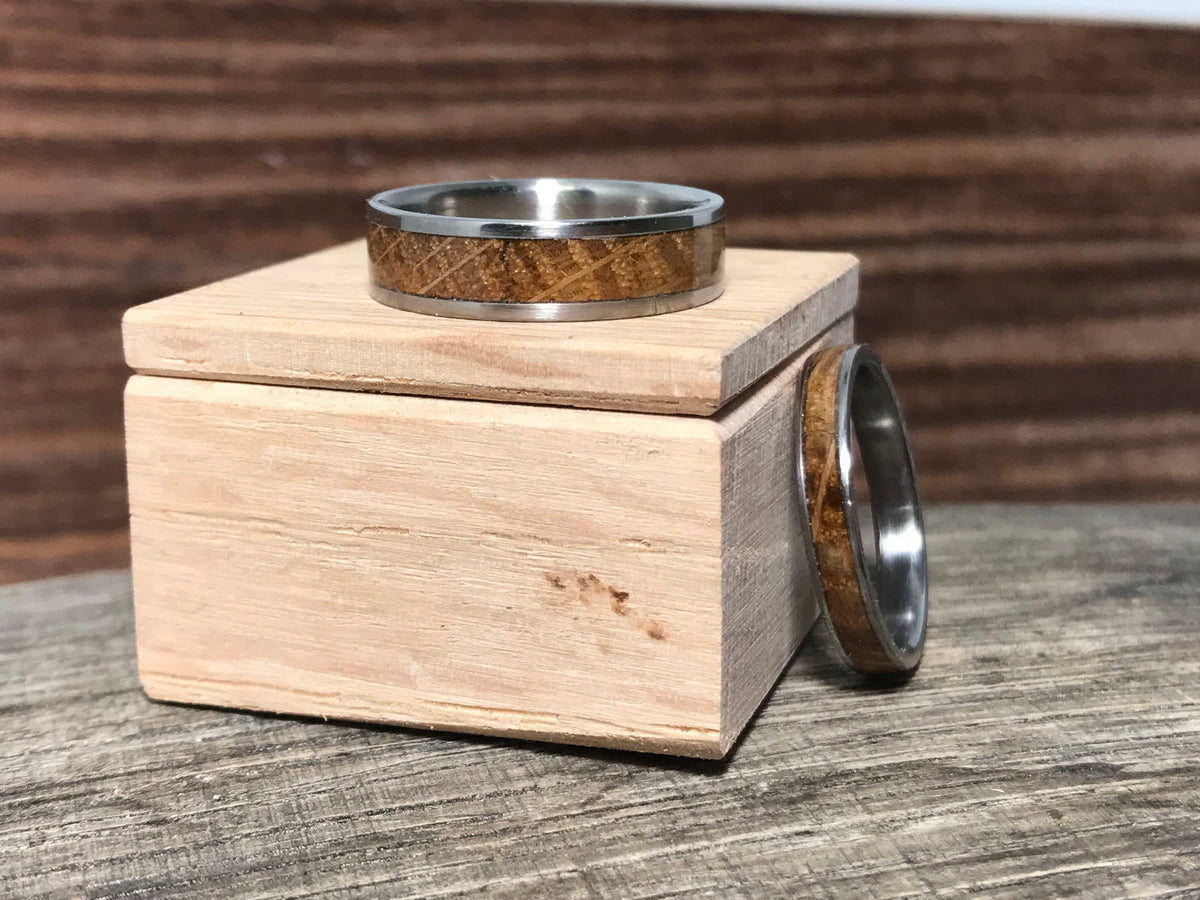 One silver-toned ring with a light brown wood inlay sits on a small light wooden box, while another ring rests on the weathered wooden surface beside it.