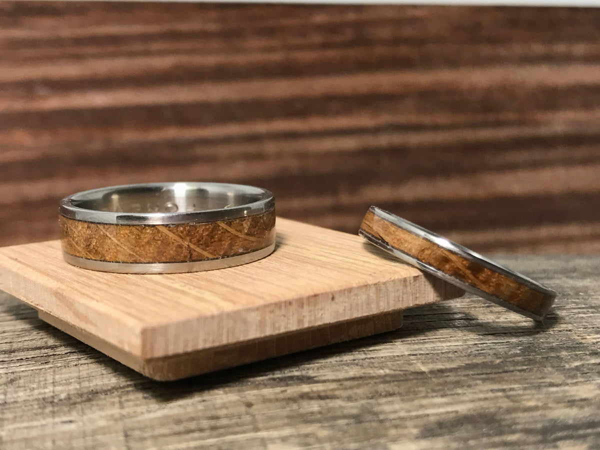 One silver-toned ring with a light brown wood inlay sits on a small light wooden platform, with another ring resting on the weathered wooden surface next to it.
