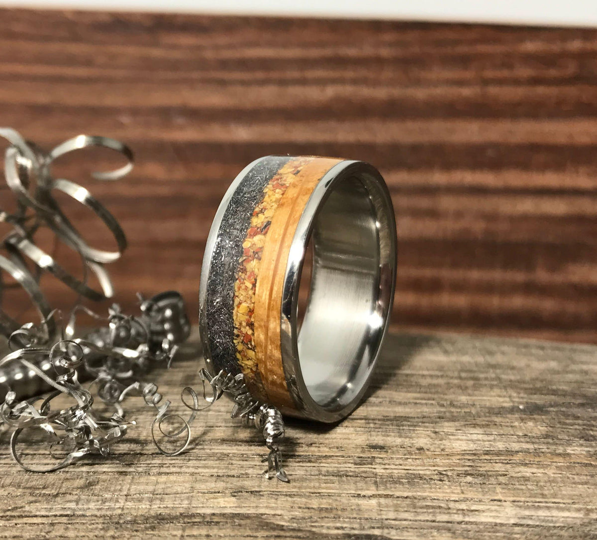 A titanium ring with a dark gray and light brown inlay is centered on a wooden surface, with metal shavings to its left.