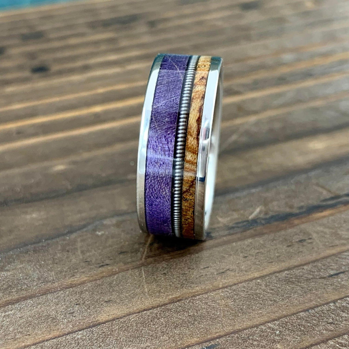 A silver ring with a purple inlay, a thin silver guitar string, and a light brown wood inlay, viewed from the side on a wooden surface.