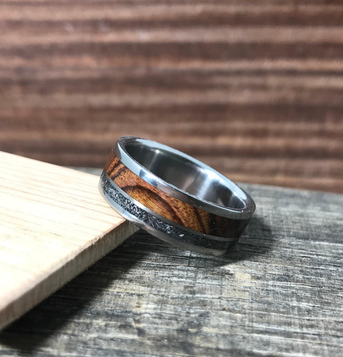 A silver-colored ring with a central wood grain band and a thin, textured gray band, positioned on a light wooden slat on a wooden surface.