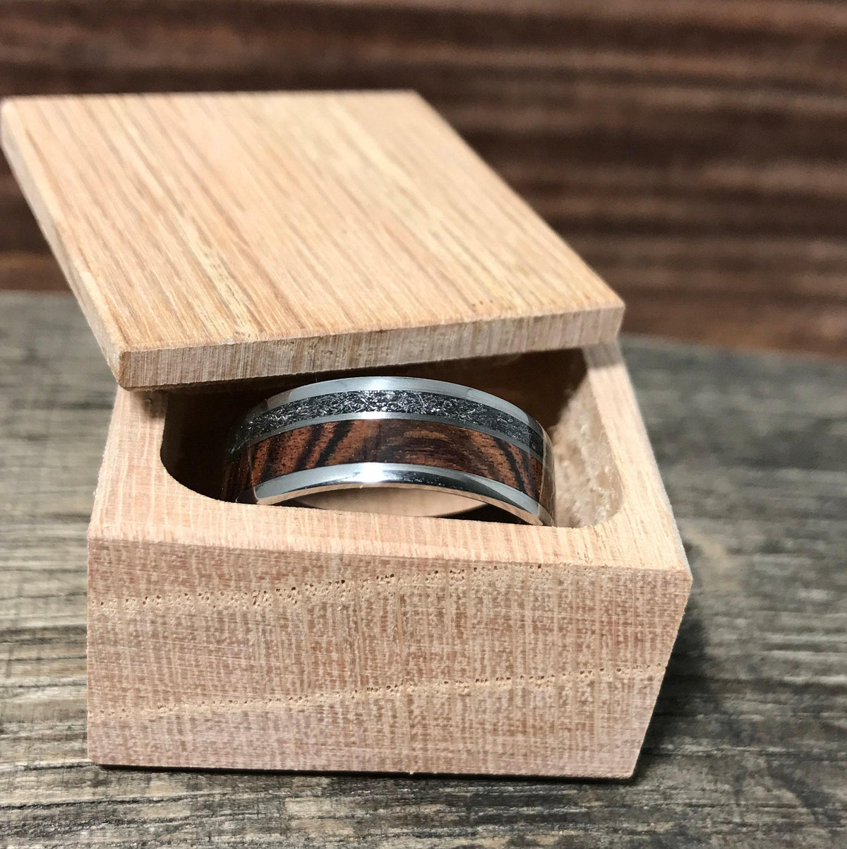A silver-colored ring with a central wood grain band and a thin, textured gray band, nestled inside an open light-colored wooden box.