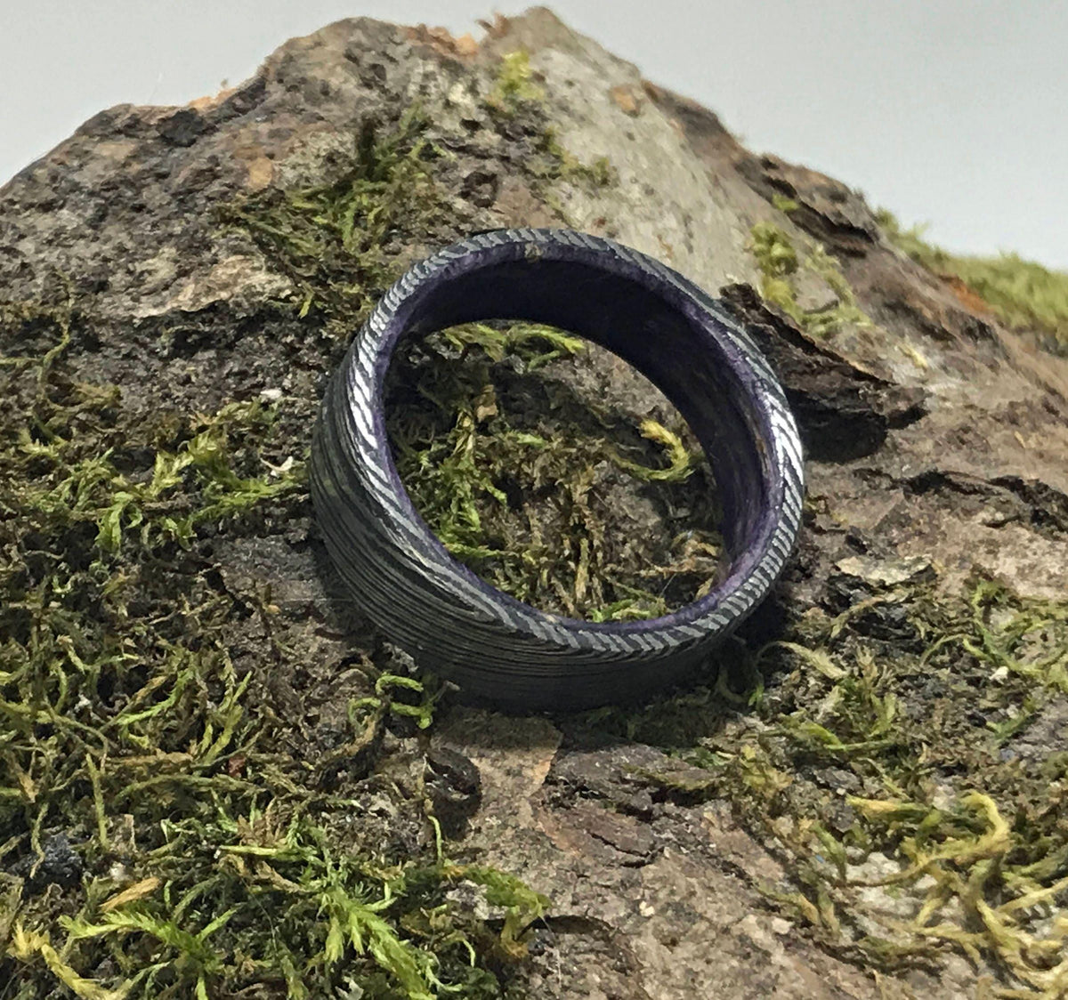 A dark gray Damascus steel ring with a purple interior is angled on a mossy log.