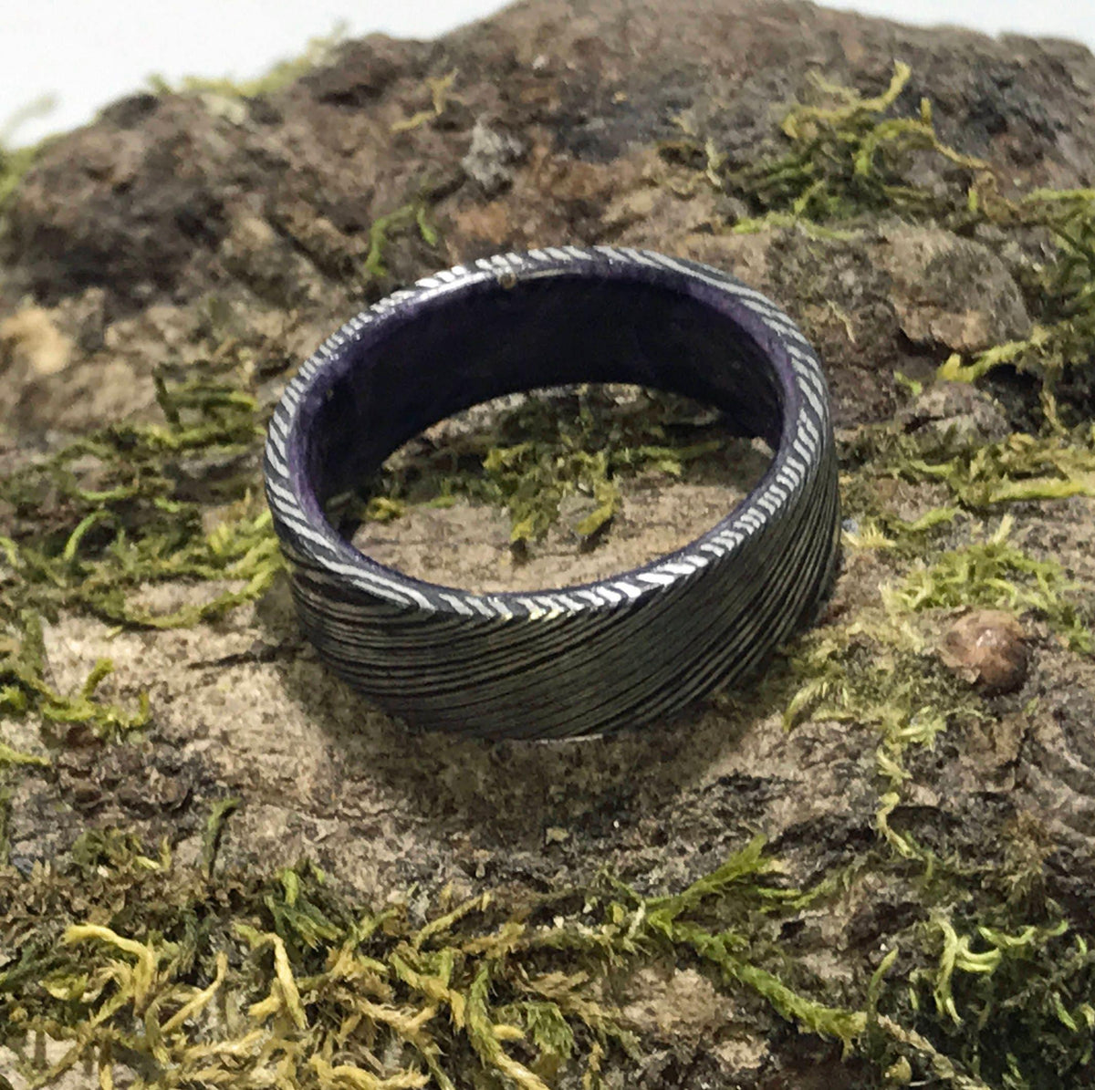 A dark gray Damascus steel ring with a purple interior is viewed from the front on a mossy log.