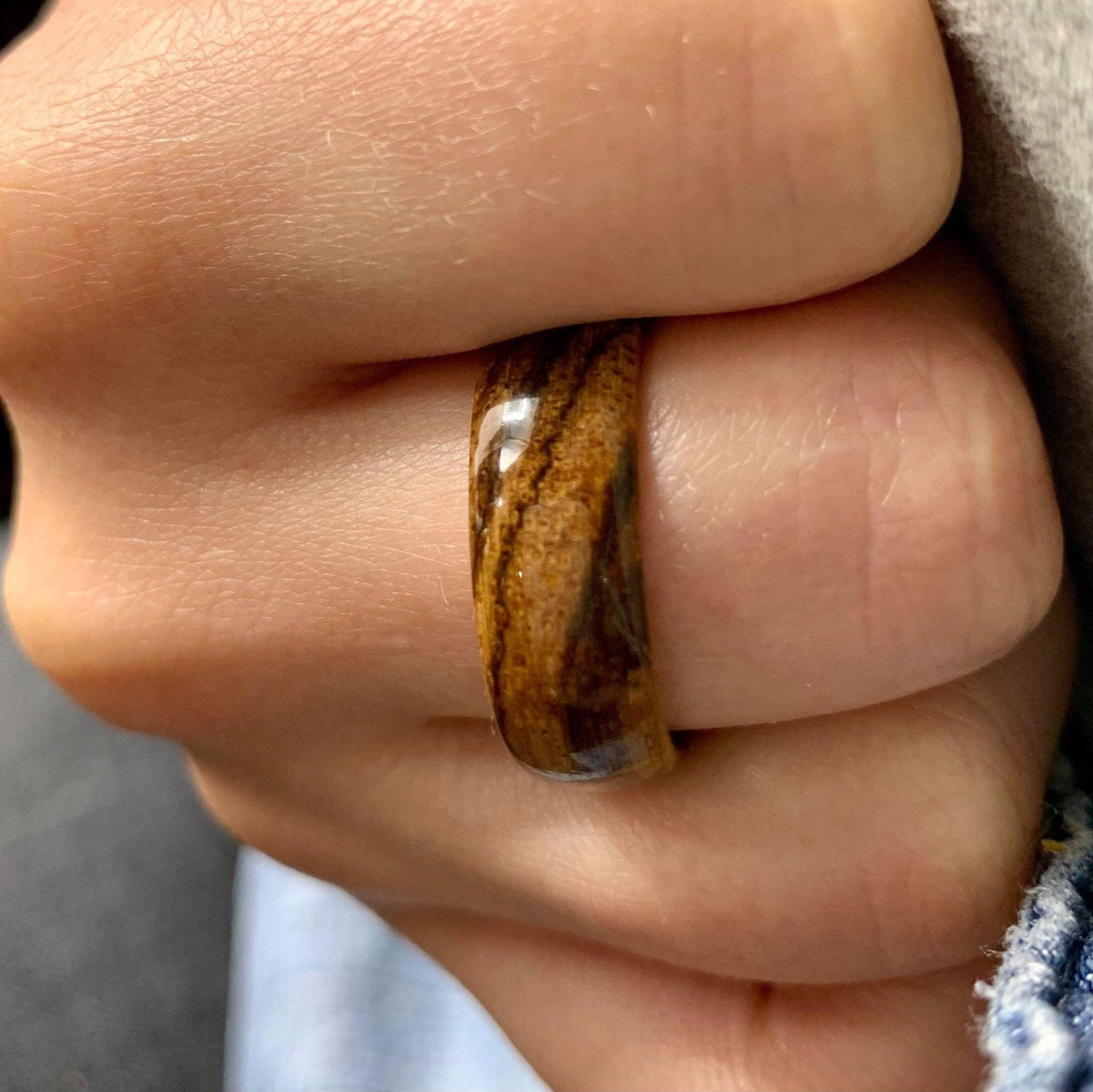 A dark brown and tan striped wooden ring is worn on a finger, with a gray sleeve and blue denim visible in the background.