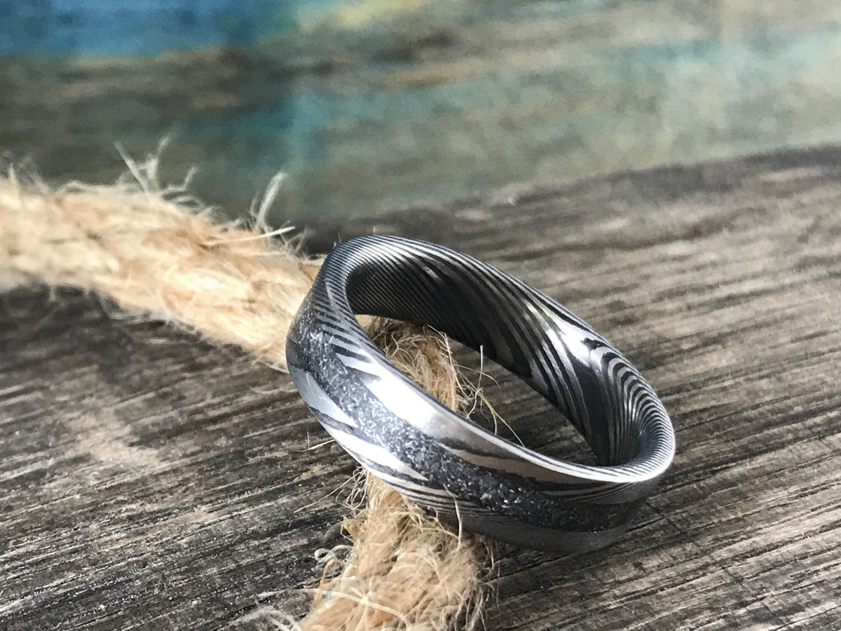 A silver-toned ring with a central textured band and patterned sides is angled on a coil of rope on a wooden surface.