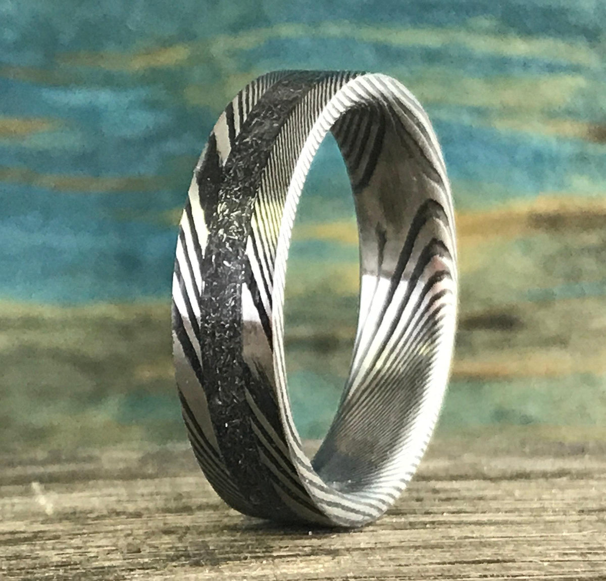 A silver-toned ring with a central textured band and patterned sides stands upright on a wooden surface.