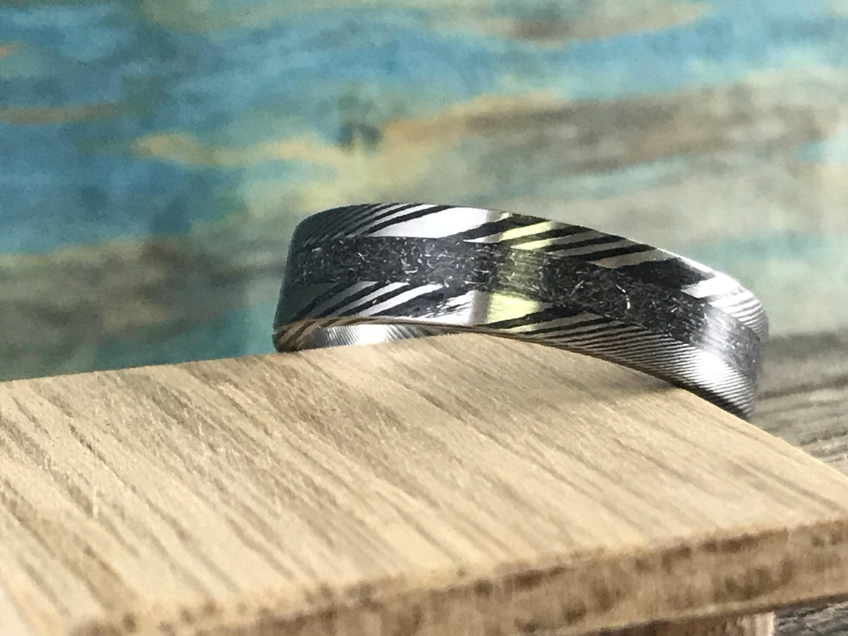 A silver-toned ring with a central textured band and patterned sides is shown in profile on a light wooden block.