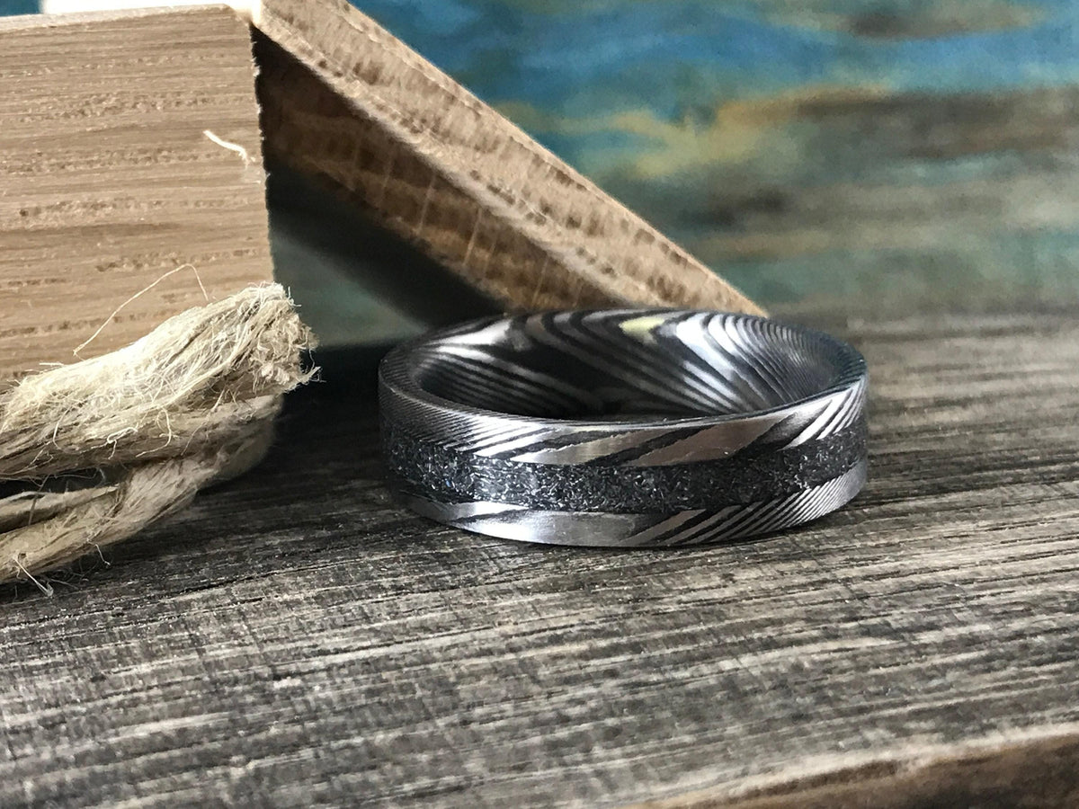 A silver-toned ring with a central textured band and patterned sides is on a wooden surface next to a rope and a wooden block.