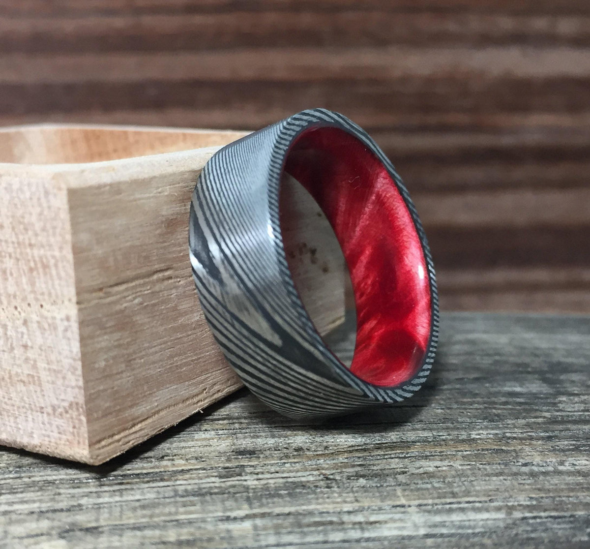 A silver-toned Damascus steel ring with a red interior, angled on a dark wooden surface with a light wooden box in the background.