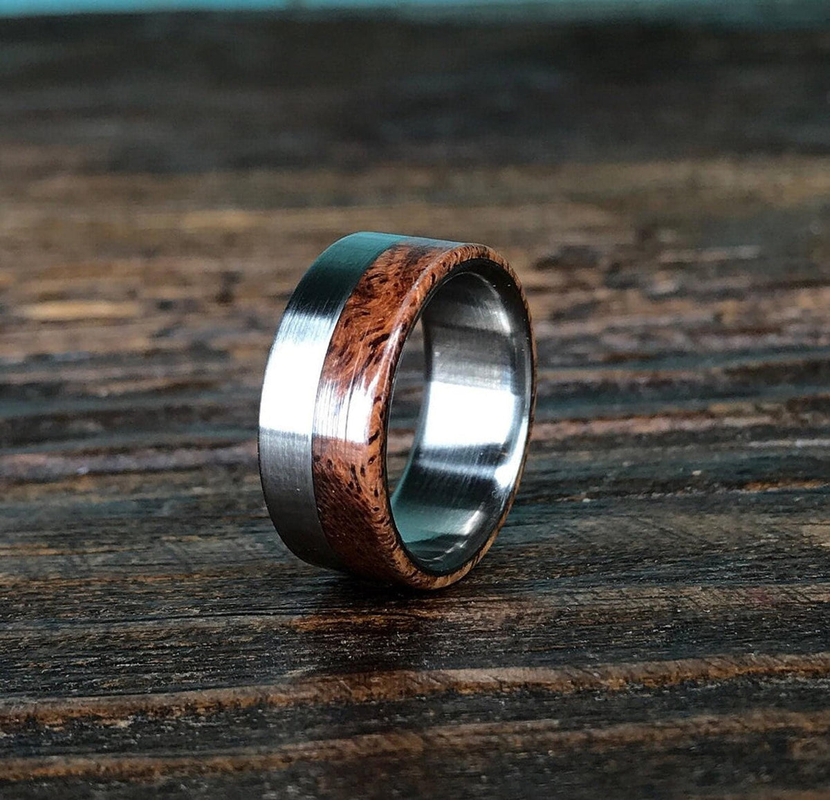 A ring with a brushed silver-colored metal band on the left, a reddish-brown wooden inlay with dark grain in the middle, and a polished silver-colored metal band on the right, on a dark wooden surface.