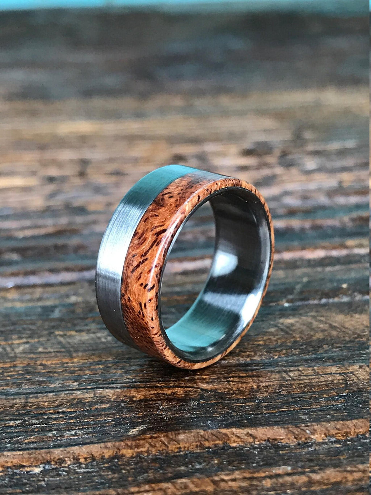 A ring with a brushed silver-colored metal band on the left, a reddish-brown wooden inlay with dark grain in the middle, and a polished silver-colored metal band on the right, on a dark wooden surface.
