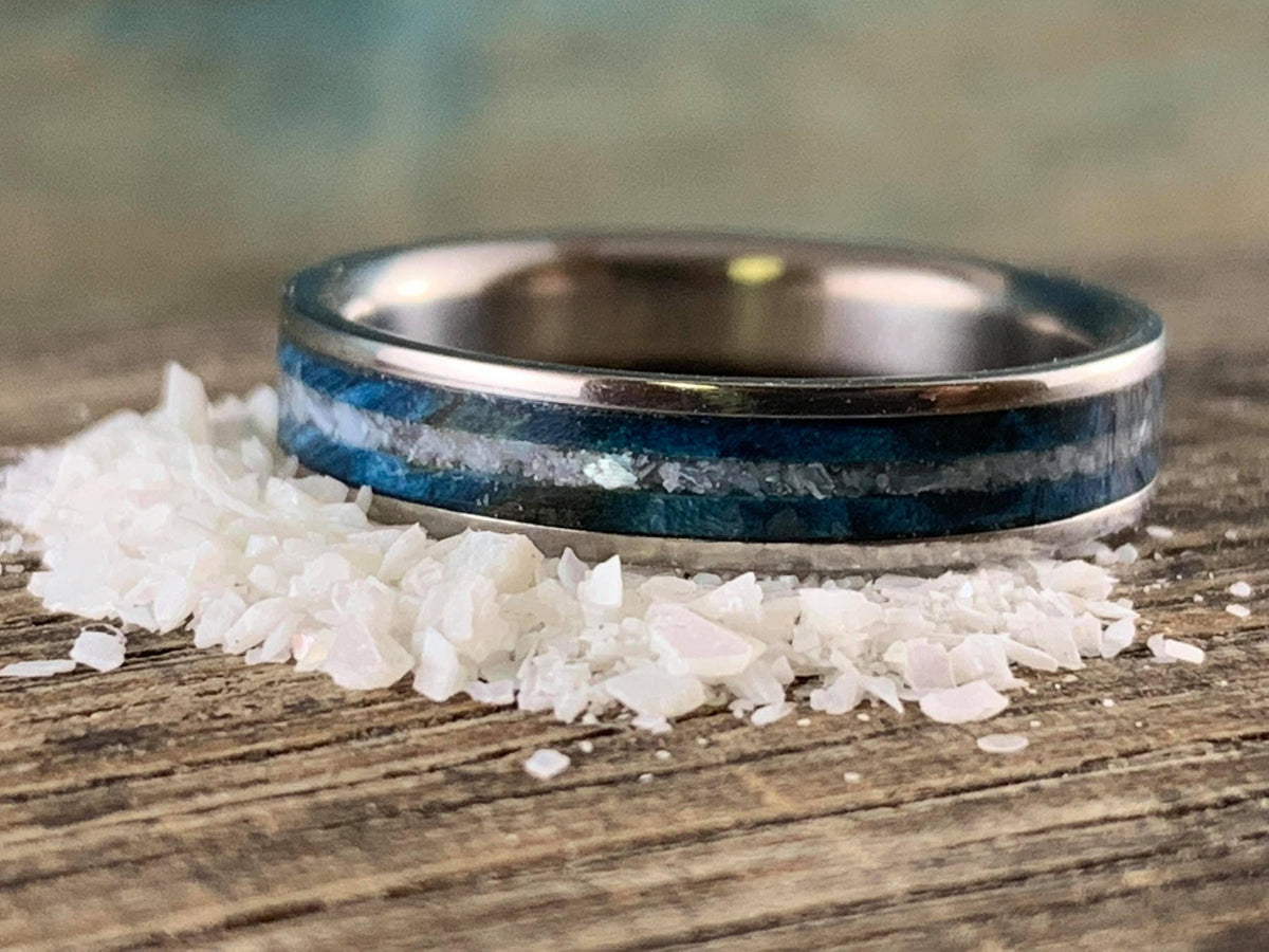 A silver-colored ring with a blue wood and white pearl inlay, resting on a pile of white crystalline flakes.