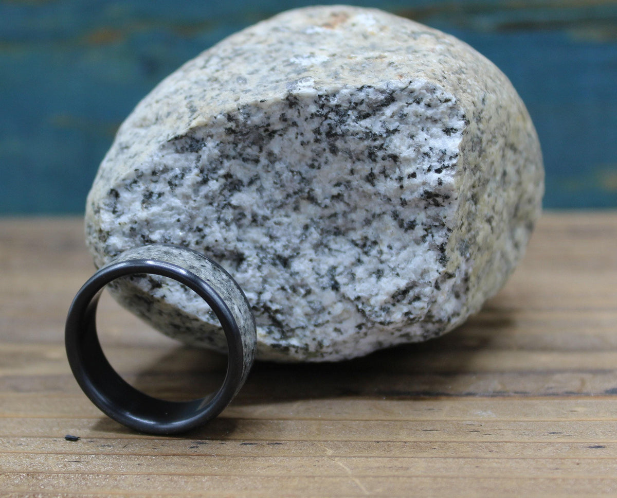 A black ring with a light gray, speckled granite inlay rests on a wooden surface in front of a large, light gray granite rock.
