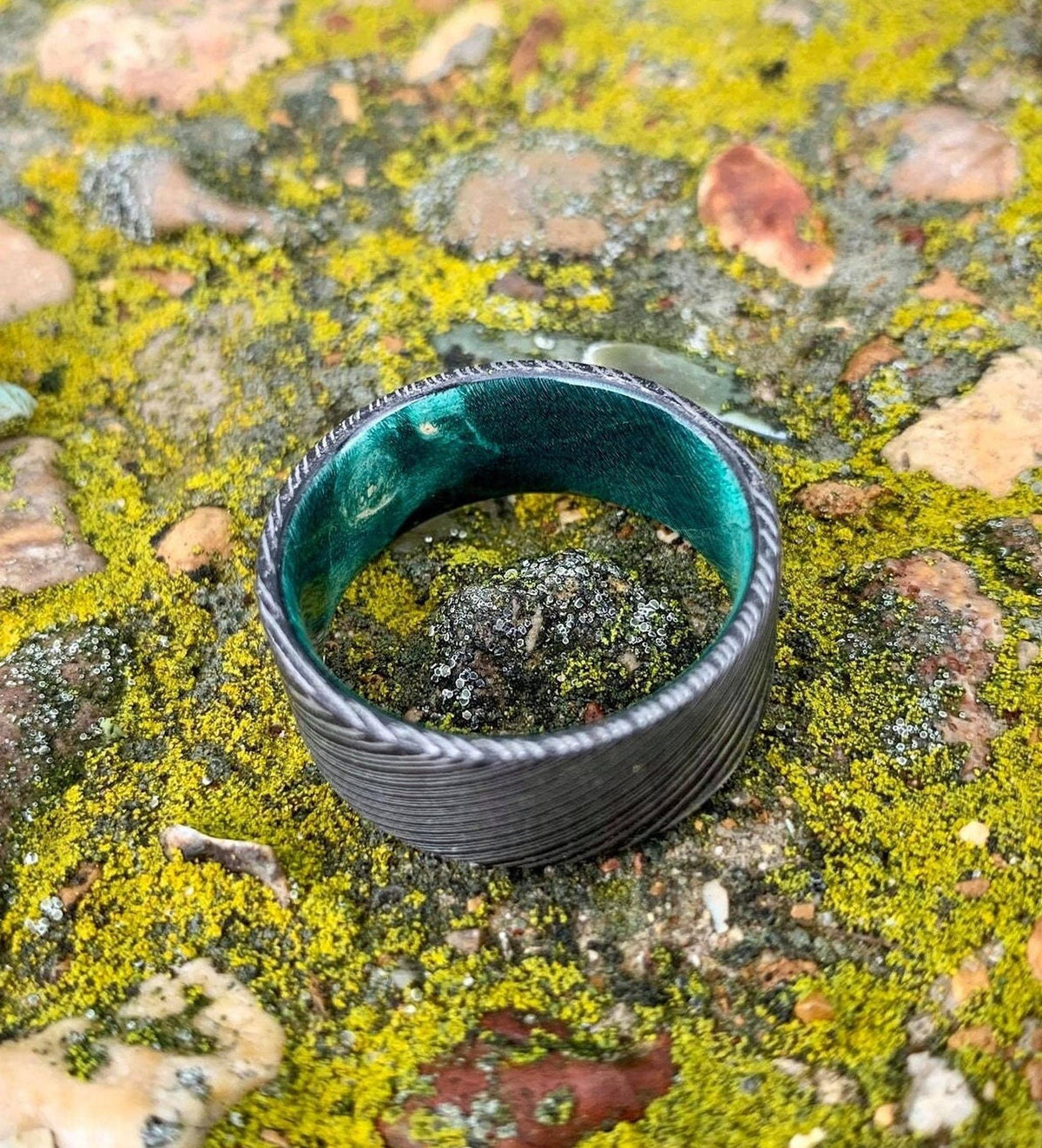 A wide, flat, patterned metal ring with a bright green interior is shown from a slightly different angle on mossy rocks.