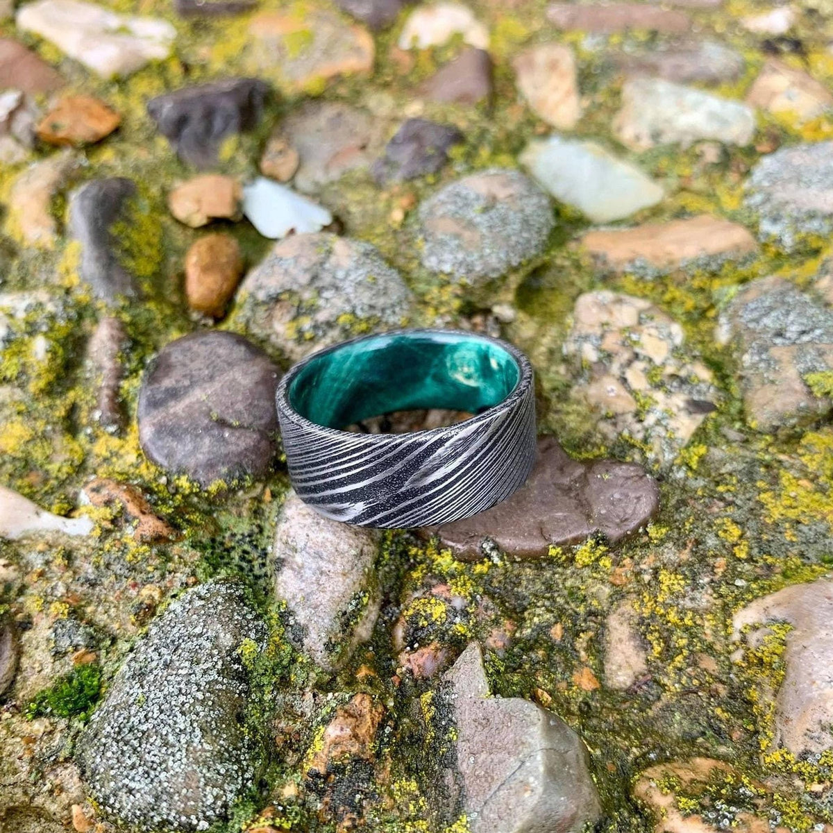 A wide, flat, patterned metal ring with a bright green interior is shown from a slightly different angle on mossy rocks, with more light on the ring.